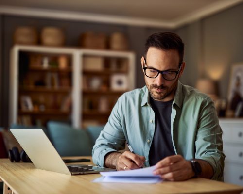 A serious, focused male freelance copywriter with reading glasses taking notes on a sheet of paper and editing his latest work.